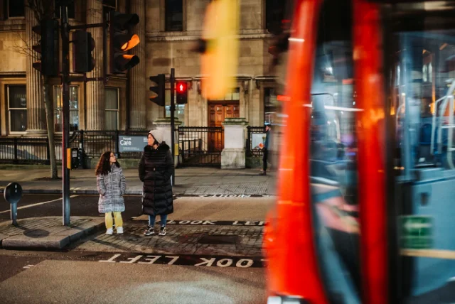 report scam text uk – woman and child waiting at a London crossing as a red bus passes, the everyday moment a scam text tends to arrive